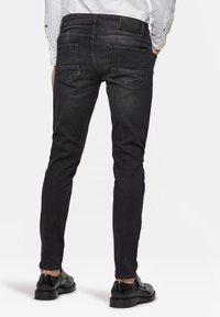 Man med slim-fit svarta jeans med bakfickor och svarta dressade skor, stående mot en enkel vit bakgrund.