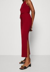 Robe sans manches bordeaux avec une taille ceinturée, une fente sur le côté et une texture lisse. Associée à des mules noires avec un accent en chaîne dorée.