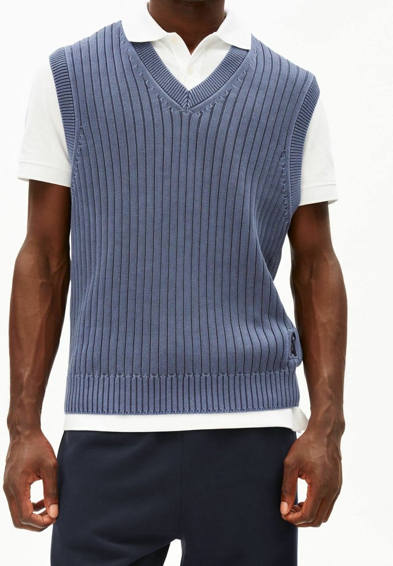 Homme portant un pull sans manches en maille côtelée bleue sur un polo blanc à manches courtes et un pantalon foncé.