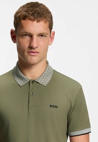 Polo de color verde oliva con cuello gris claro estampado, mangas cortas y diseño ajustado que presenta un logo negro en el pecho izquierdo.