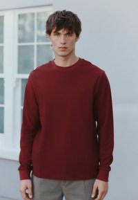 Langærmet, ribbet burgunderrød skjorte, rund halsudskæring, blødt stof. Båret med lysegrå bukser, model vender fremad, minimalistisk baggrund.