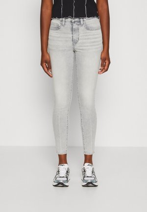 Personne portant un jean skinny gris clair, des baskets blanches et grises, et un top court noir, debout devant un fond blanc uni.