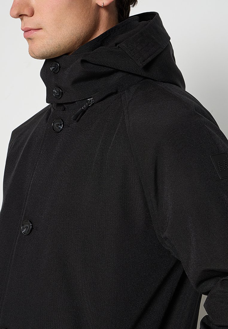 Zwarte hoodiejas met een gestructureerde afwerking, voorzien van grote knopen, een ritszak en een logo patch op de mouw.