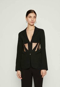 Uma jovem com cabelo para trás usa um blazer preto com recortes transparentes na cintura, combinado com calças pretas e brincos de argola.