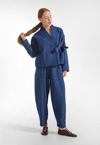 Kobieta z rudymi włosami związanymi w kucyk, ubrana w niebieską pikowaną kurteczkę zapinaną na rzep i pasujące luźne spodnie, stoi na białym tle, poprawiając włosy.