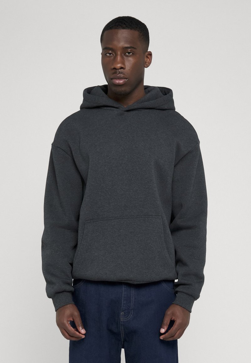 Jeune homme noir portant un sweat à capuche gris foncé et un pantalon bleu foncé, debout devant un fond gris clair uni.