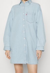 Abito camicia in denim azzurro chiaro con maniche lunghe, chiusura a bottoni sul davanti, taschino sul petto e vestibilità leggermente oversize. Tessuto morbido e design casual.
