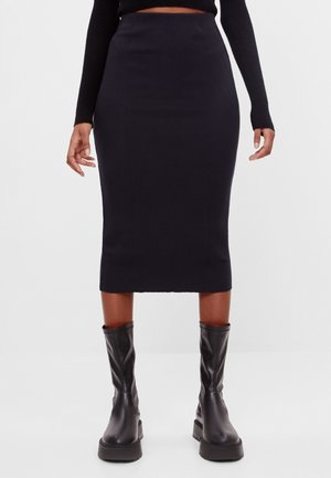 Personne portant une jupe midi noire ajustée, un haut noir à manches longues et des bottes en cuir noires mi-mollet, debout, les bras détendus.