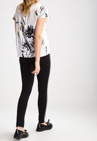 Donna che indossa una camicia bianca a maniche corte con fantasia floreale, pantaloni neri slim con striscia laterale e scarpe nere a lacci, in piedi davanti a uno sfondo neutro.