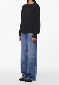 Černá oversized mikina s dlouhými rukávy, která je kombinována se světle modrými širokými džíny a černými botami, proti bílému pozadí.