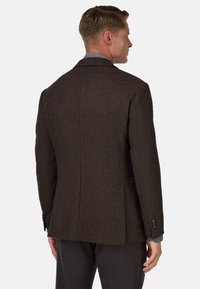 Brązowy żakiet w kratę houndstooth o teksturowanym wykończeniu, z pojedynczym rozcięciem z tyłu, dwoma guzikami na rękawach i kieszeniami bocznymi. Pod spodem jest jasnoszara koszula.