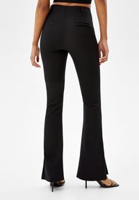 Kvinna i svarta högmidjade flarebyxor och svarta öppna klackar, stående med avslappnade armar vid sidorna mot vit bakgrund.