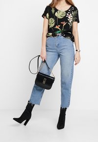 Top nero a maniche corte con stampa floreale, jeans blu a vita alta, stivali con tacco neri e una borsa nera con tracolla a catena.