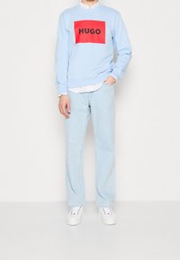 Sweat-shirt bleu clair avec un logo rectangulaire rouge portant l'inscription "HUGO", porté sur une chemise blanche et associé à un jean bleu clair et des baskets blanches.