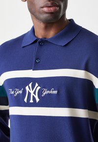 Temno modra polo majica iz bombaža, opremljena z belimi črtami in logotipom. Ima tradicionalen ovratnik in gumbasto zapenjanje.