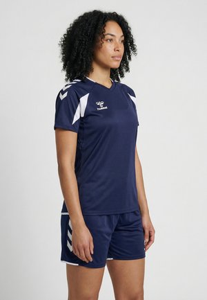 Vrouw die een donkerblauw-wit sportshirt en korte broek draagt, in profiel staand tegen een effen lichte achtergrond.