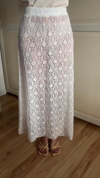Falda maxi blanca de encaje con estampado floral, cintura elástica, tela semitranslúcida, combinada con chanclas amarillas sobre un suelo de madera.