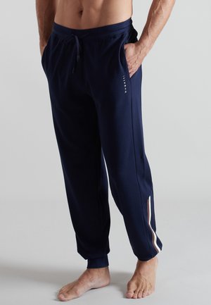 Mann trägt marineblaue Jogginghosen mit Seitentaschen, einem kleinen Logo am Oberschenkel und beigen sowie weißen Seitenstreifen, steht barfuß.