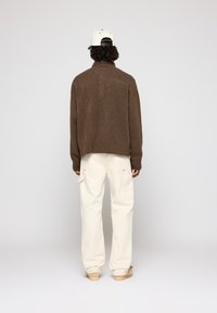 Suéter castanho tricotado com gola alta, combinado com calças largas brancas. O conjunto apresenta um design minimalista e uma textura suave.