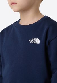 Marineblå genser laget av mykt stoff, med rund hals og hvit logo på brystet. Minimalistisk design med jevn tekstur.