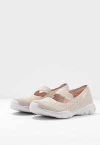 Zapatos de punto beige con una correa elástica tipo Mary Jane, parte superior texturizada y suela de goma blanca. Diseño abierto con tela transpirable.