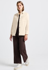 Kremowa kurtka zapinana na guziki z kołnierzykiem i dwiema kieszeniami na piersiach, w połączeniu z brązowymi spodniami o szerokich nogawkach oraz białymi trampkami. Gładka faktura.