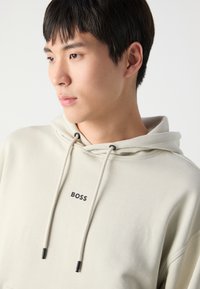 Sudadera beige con cordones y un logo frontal "BOSS." La tela parece suave con una textura lisa y un diseño de corte relajado.