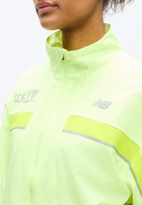 Κοντινό πλάνο ενός ατόμου που φοράει κίτρινο μπουφάν με φερμουάρ υψηλής ορατότητας, με αντανακλαστικές λωρίδες και λογότυπο New Balance στο στήθος.