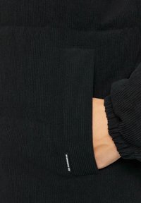 Zwarte pullover met ribbeltextuur en elastische boorden. Voorzien van een zijzak en een merklabel aan de onderrand. Zacht materiaal.