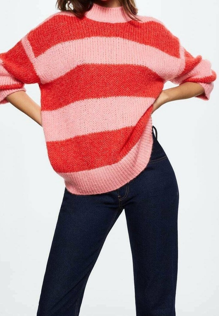 Person, der bærer en kraftig strikket sweater med brede pink og røde vandrette striber og mørkeblå jeans, hænderne hvilende på hoften.