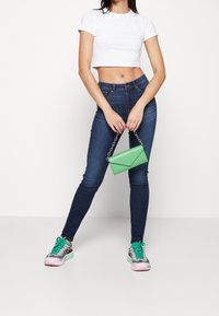 Mujer con una camiseta blanca recortada y jeans oscuros ajustados sostiene un bolso verde, vistiendo zapatillas rosas y verdes, de pie frente a un fondo blanco.