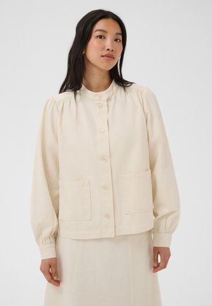 Femme aux longs cheveux foncés portant une veste crème à boutons avec des poches avant et une jupe assortie, sur un fond blanc uni.