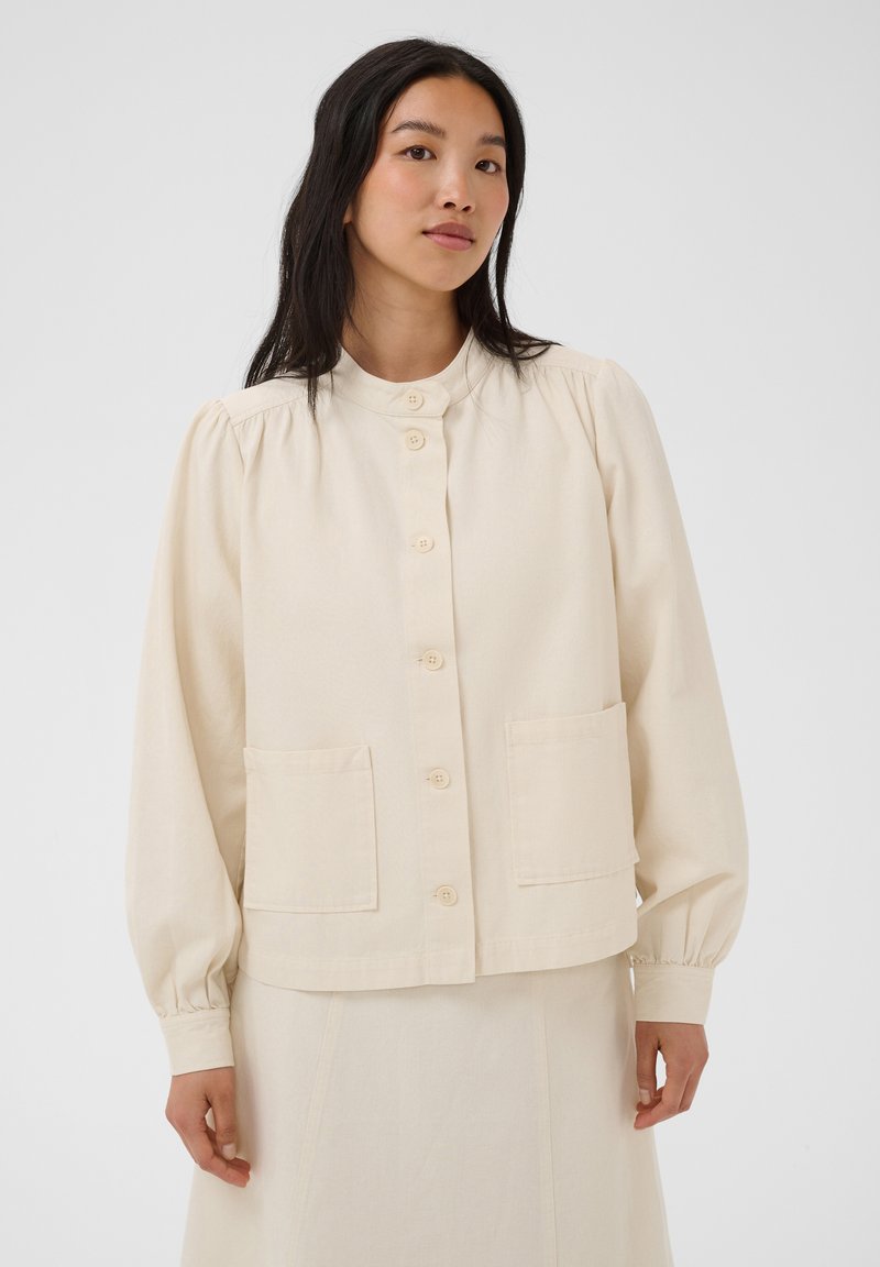 Femme aux longs cheveux foncés portant une veste crème à boutons avec des poches avant et une jupe assortie, sur un fond blanc uni.