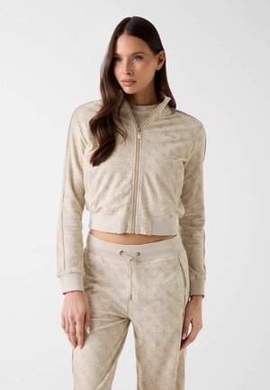 Femme aux longs cheveux bruns portant une veste courte beige à motifs et fermeture éclair, ainsi qu’un pantalon assorti à cordon, sur fond uni.