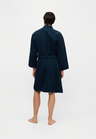 Homme debout pieds nus portant un peignoir bleu foncé texturé avec une ceinture, tourné de dos sur un fond clair uni.