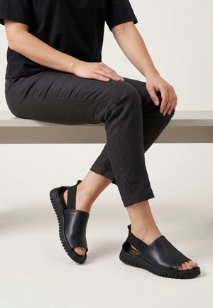 Persoon zit op een bankje, draagt zwarte open teen instapschoenen met dikke zolen en elastische hielbandjes, gekleed in een zwart top en donkere broek.