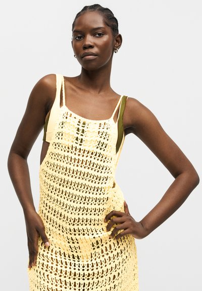 Γυναίκα με κοτσίδες, που φοράει κίτρινο πλεκτό αμάνικο φόρεμα πάνω από ένα ελιά πράσινο τοπ, ποζάρει με τα χέρια στη μέση μπροστά από ένα απλό φόντο.