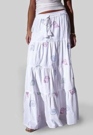 Witte maxirok met lagen en meerkleurige schelpenprints, taille met aantrekkoord en kwastjes, gecombineerd met een wit topje en een gouden armband.