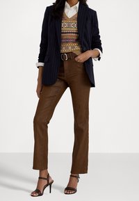 Femme portant un pantalon taille haute marron, un gilet en tricot à motifs colorés, une chemise blanche, un blazer bleu marine à fines rayures et des sandales à talons marron foncé.