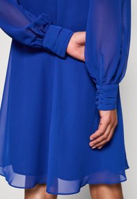 Femme portant une robe bleu roi avec des manches longues transparentes et des poignets boutonnés, une main reposant derrière le dos.