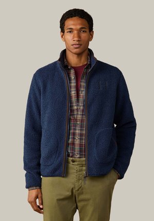 Mann trägt eine blaue Fleecejacke mit Reißverschluss über einem rot-grün karierten Hemd und olivgrünen Hosen, steht vor einem schlichten beigen Hintergrund.