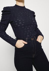 Námořnická modrá dlouhá rukávová blůza s nabíranými rameny, která má diamantový vzor a průhledné části. Kombinováno s džínami s vysokým pasem v tmavé barvě.