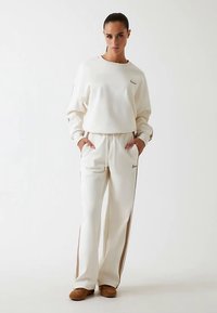 Felpa bianca e pantaloni della tuta abbinati con strisce laterali marroni. Il tessuto sembra morbido; i pantaloni hanno tasche. Il modello indossa pantofole marroni.