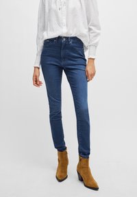Smala blå denimjeans med fem fickor, medelhög midja och en slät textur, kombinerade med tanmocka ankelboots och vit blus.