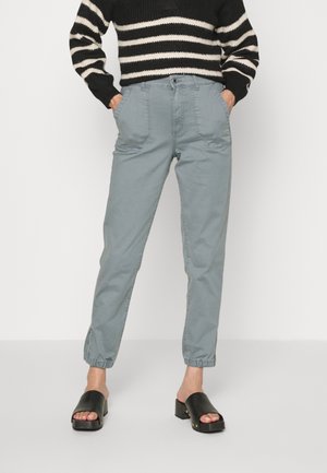 Person trägt hellblaue, zulaufende Hosen mit elastischen Bündchen, schwarz-weiß gestreiften Pullover und schwarze Sandalen mit offenem Zehenbereich und Blockabsatz.