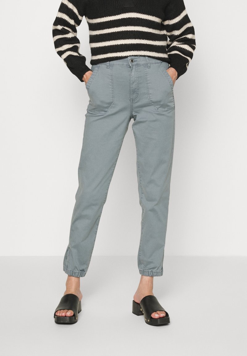 Personne portant un pantalon bleu clair fuselé avec des poignets élastiques, un pull rayé noir et blanc, et des sandales noires à talon bloc et bout ouvert.