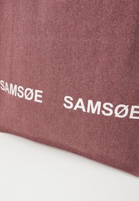 Mørkerød tekstilpose med merkevarenavnet "SAM SøE" i hvite bokstaver. Glatt tekstur med synlige vevde fibre. Enkel design.