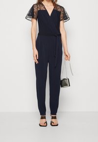 Combinaison cache-cœur bleu marine avec manches courtes en dentelle, taille ceinturée, et pantalons fuselés. Portée avec des sandales à talons noires et un petit sac à main.