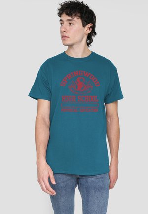 Young man with curly dark hair wearing a teal T-shirt with red "Springwood High School Physical Education" text and blue jeans, looking left.