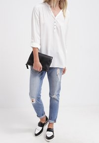 Blouse blanche à manches longues avec un col boutonné, associée à un jean déchiré en teinte claire et des mocassins bicolores noir et blanc. Pochette noire à la main.